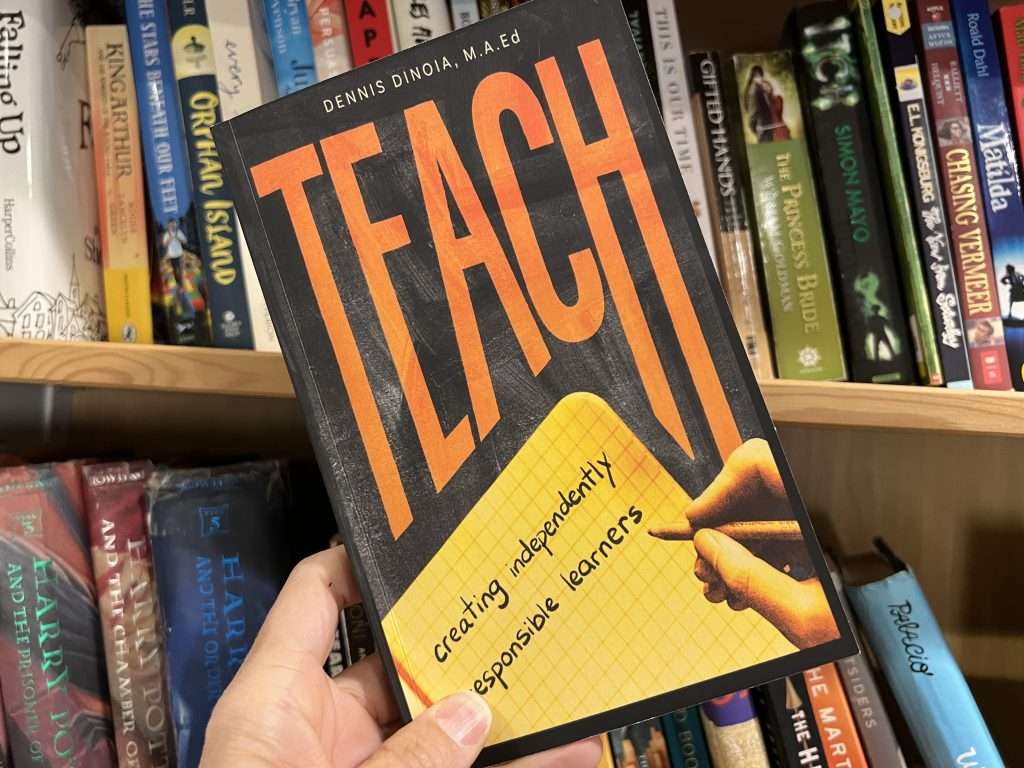 Teach: Creativing Independently Responsible Learners by Dennis DiNoia held in the air wiht a bookshelf background.